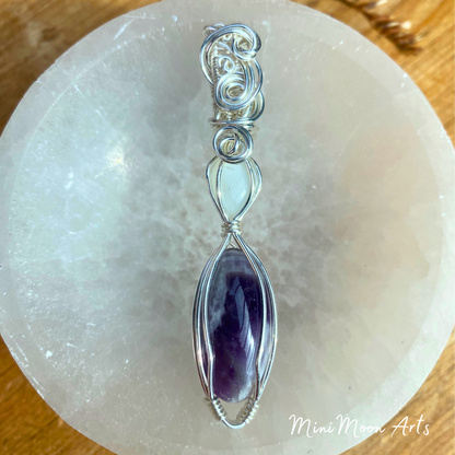 moonstone and amethyst wire wrapped silver wire pendant sat at a front view angle in a selenite bowl with a wooden table back ground